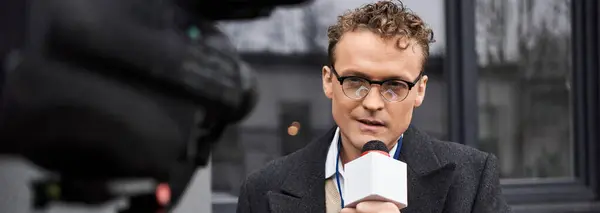 Il giornalista, indossando occhiali e un cappotto, trasmette commenti penetranti in un microfono all'aperto. — Foto stock