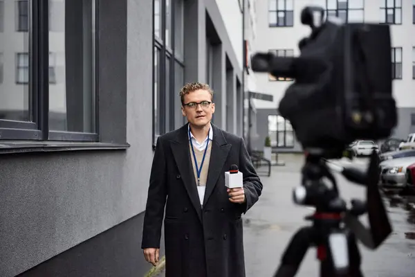 Ein engagierter Nachrichtensprecher interviewt draußen, hält ein Mikrofon in der Hand, während er Mantel und Brille trägt. — Stockfoto