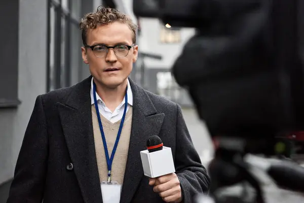 Un giornalista concentrato con occhiali e un cappotto riporta aggiornamenti mentre tiene un microfono all'aperto. — Foto stock