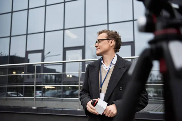 A journalist wearing glasses and a coat engages with a microphone outside an office building, sharing updates. — Stock Photo