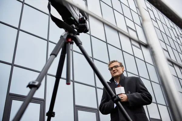 Un periodista con gafas y un abrigo habla en un micrófono en un lugar contemporáneo durante el día. - foto de stock