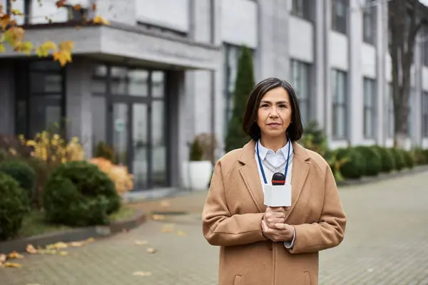 Una donna multirazziale in un cappotto beige si erge con fiducia con un microfono, riferendo sul posto. — Foto stock