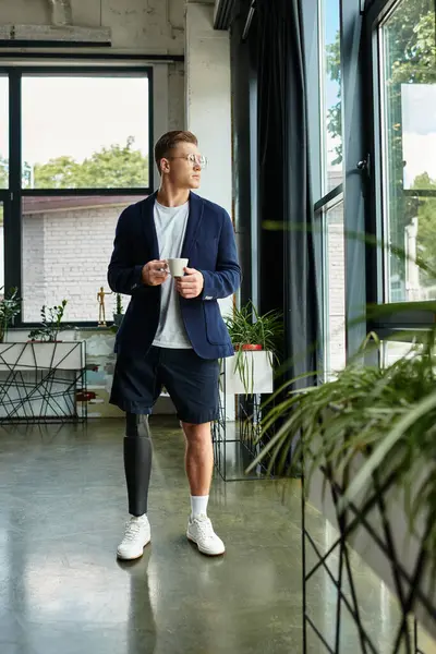 Um jovem empresário fica ao lado da janela tomando café, mostrando resiliência e estilo em um espaço de trabalho moderno. — Fotografia de Stock