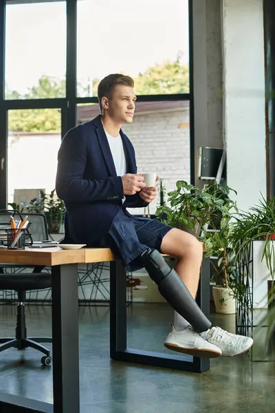 Un joven hombre de negocios con un miembro artificial se sienta con confianza en un escritorio, bebiendo café en una oficina elegante. - foto de stock