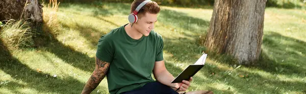 Un joven guapo se relaja al aire libre, inmerso en un libro mientras usa auriculares. - foto de stock