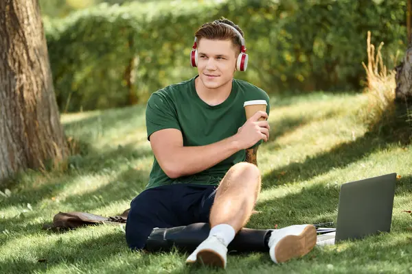 A young man relaxes on the grass, sipping coffee while working on his laptop, embodying inclusion and joy. — Stock Photo