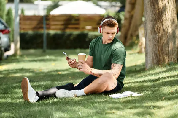 A young man with a prosthetic leg sits comfortably on the grass, sipping coffee and listening to music. — Stock Photo