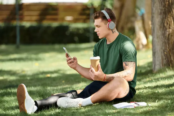 A young man with a prosthetic leg relaxes on the grass, enjoying coffee, music, and his phone. — Stock Photo