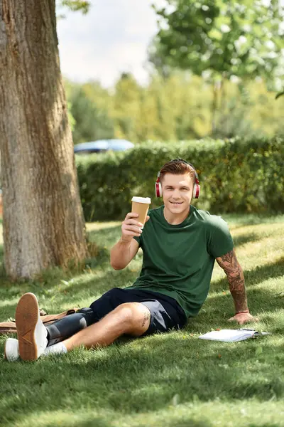 A young man with a prosthetic leg relaxes on the grass, wearing headphones and sipping a drink with joy. — Stock Photo