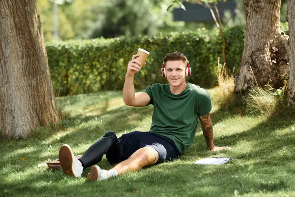 Un joven alegre con una pierna protésica se sienta en la hierba, tomando una copa y disfrutando del aire libre. - foto de stock