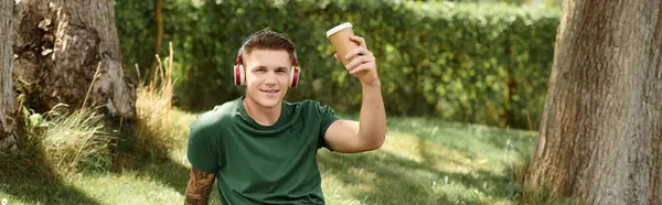 Un giovane uomo siede sull'erba, indossa le cuffie e sorride mentre tiene in mano una tazza di caffè, abbracciando la vita e la gioia. — Foto stock