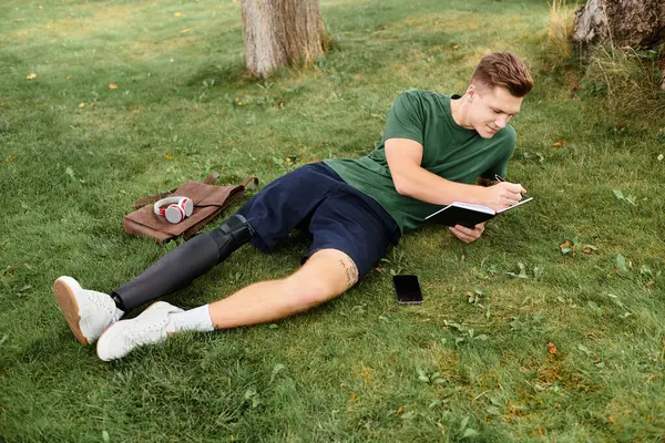 A young man with a prosthetic leg sits on grass, sketching in a notebook while relaxed and smiling. — Stock Photo