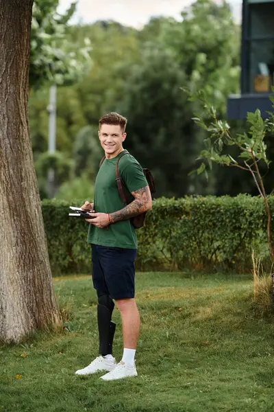 A handsome man with a prosthetic leg stands in a green park, smiling and taking notes as he embraces nature. — Stock Photo