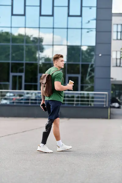 A young man with a prosthetic leg strides confidently outdoors, carrying a coffee in one hand. — Stock Photo