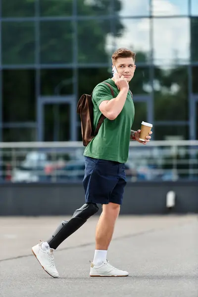 Un hombre joven y guapo con una pierna protésica camina afuera, charlando en su teléfono y sosteniendo una taza de café. - foto de stock