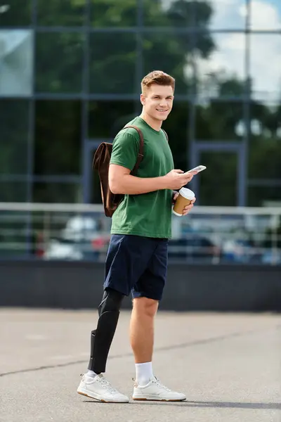 Ein junger, gutaussehender Mann mit Beinprothese steht draußen, lächelt und nippt an seinem Smartphone, während er Kaffee schlürft.. — Stockfoto