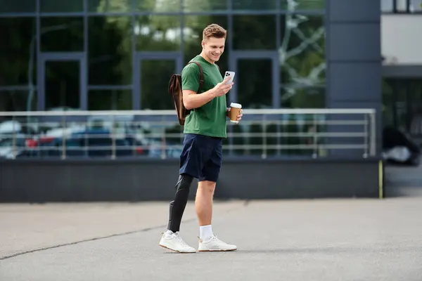 Un jeune homme souriant avec une jambe prothétique se tient à l'extérieur, engageant avec son téléphone et savourant du café. — Photo de stock