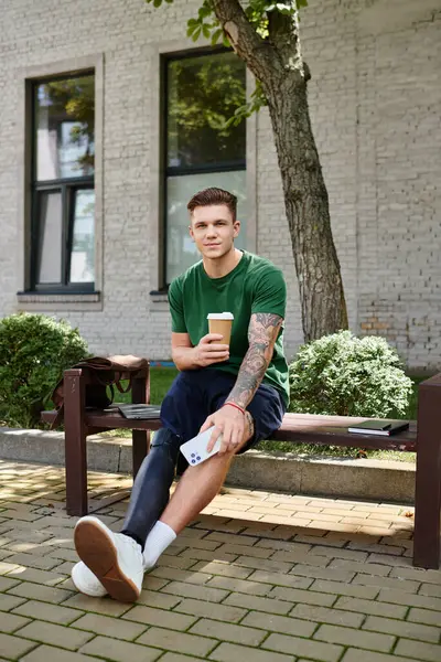 A young man with a prosthetic leg relaxes on a bench, sipping coffee in a vibrant city park filled with greenery. — Stock Photo