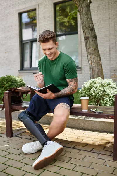 Ein junger, gutaussehender Mann mit Beinprothese sitzt auf einer Bank und lächelt, als er in ein Notizbuch schreibt.. — Stockfoto