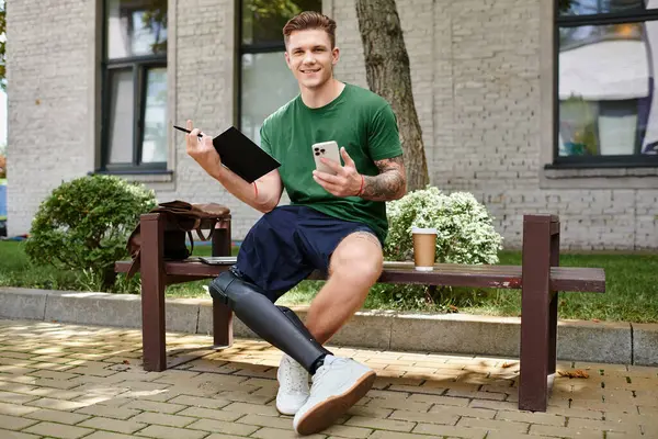 A young man sits on a bench outside, smiling as he balances a notebook and phone, expressing joy and creativity. — Stock Photo