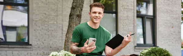 A young man sits comfortably outdoors, smiling as he writes in a notebook while using his phone. — Stock Photo