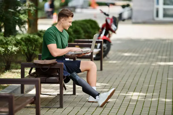 Um jovem com uma perna protética se senta ao ar livre, focado em seu laptop em meio a vegetação exuberante e vida urbana. — Fotografia de Stock