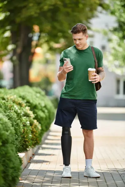 A young man with a prosthetic leg sips coffee while checking his phone, enjoying a sunny day outdoors. — Stock Photo