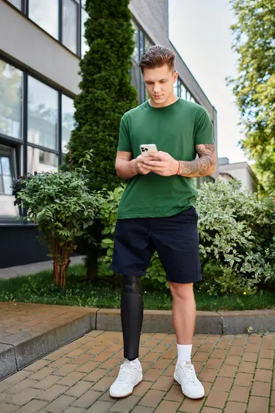 Ein junger Mann mit Beinprothese steht draußen und schreibt bequem SMS auf seinem Handy in einer lebhaften Umgebung. — Stockfoto