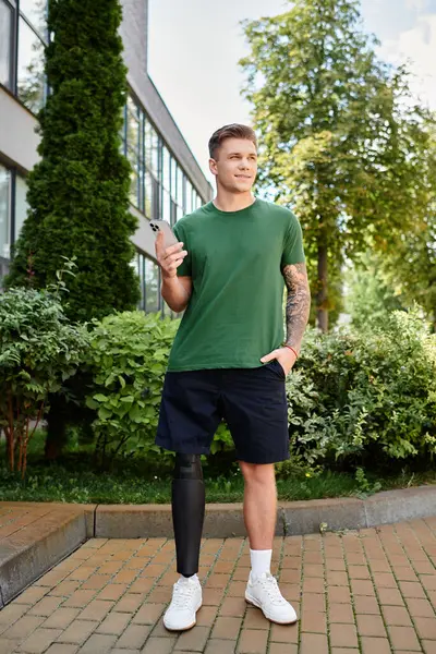 A young and handsome man confidently stands outdoors, showcasing his prosthetic leg while holding a smartphone. — Stock Photo