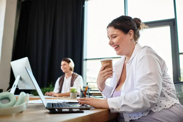 A young plus size woman smiles as she types on her laptop, sipping coffee in a cozy workspace. — Stock Photo