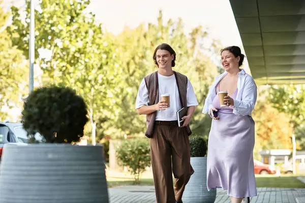 A young plus size woman and her male friend walk together, enjoying their drinks under the sun. — Stock Photo