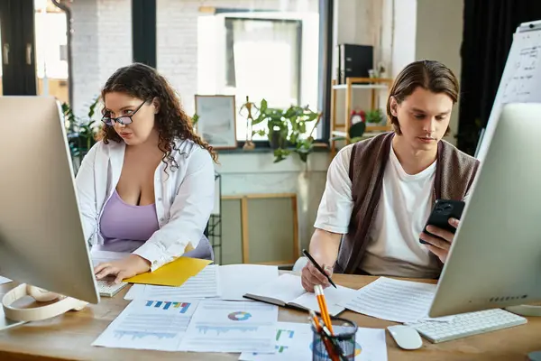 A young plus size woman focuses intently on her computer while her friend checks his phone. — Stock Photo