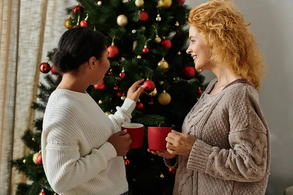 Dois parceiros felizes admiram ornamentos em sua árvore festiva enquanto desfrutam de chá quente juntos. — Fotografia de Stock