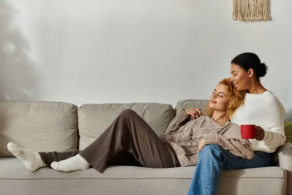 Dos mujeres se relajan juntas en su acogedora casa, compartiendo el amor y una bebida caliente en el sofá. — Stock Photo