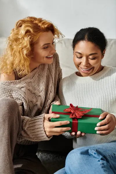 Two women express happiness while exchanging a wrapped gift in their cozy living space. — Stock Photo