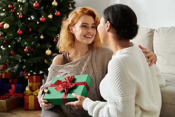 A joyful couple exchanges gifts in their cozy, holiday decorated living room. — Stock Photo