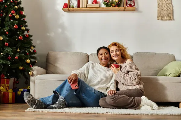 A happy couple sits on the floor, sharing a warm drink and laughter in their festive living room. — Stock Photo