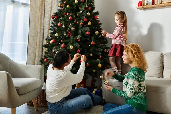 A loving lesbian couple and their daughter joyfully decorate a Christmas tree together. — Stock Photo