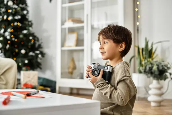 A cheerful Asian boy standing near decorated Christmas tree, holding his camera — Stock Photo
