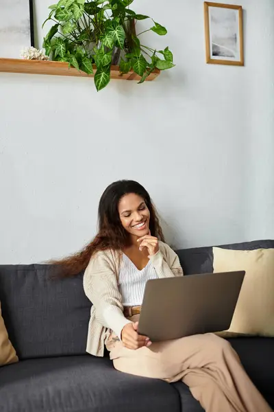 A cheerful woman sits on a soft couch, smiling while using her laptop in a warm and inviting space. — Stock Photo