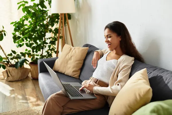 A cheerful woman sits on a sofa, happily engaged with her laptop in a bright, inviting space. — Stock Photo
