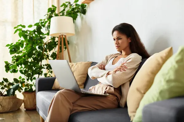 A woman sits thoughtfully on a couch, engaging with her laptop amid vibrant indoor plants. — Stock Photo