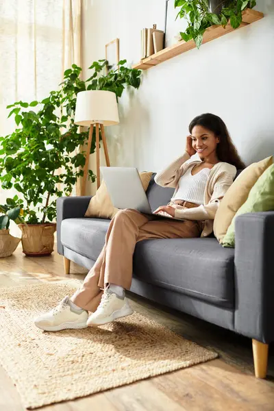 Une femme se détend sur un canapé confortable, engagée dans son ordinateur portable que la lumière du soleil filtre à travers. — Photo de stock