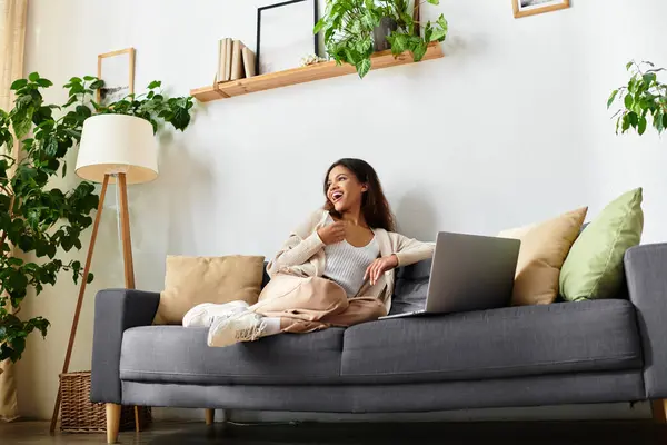 A cheerful woman lounges on a stylish sofa, engaging with her laptop in a lively environment. — Stock Photo