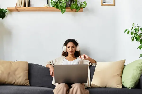 Uma jovem se senta confortavelmente em um sofá, profundamente focada em seu laptop com fones de ouvido em. — Fotografia de Stock