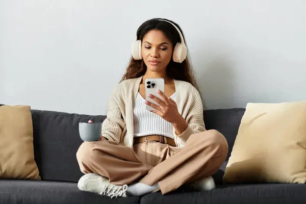 A woman with headphones sits comfortably, scrolling through her phone while sipping a warm drink. — Stock Photo