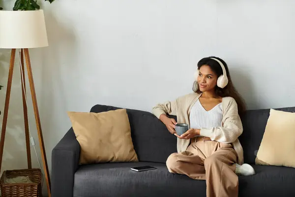 A woman sits comfortably on a couch, sipping tea while listening to music through headphones. — Stock Photo