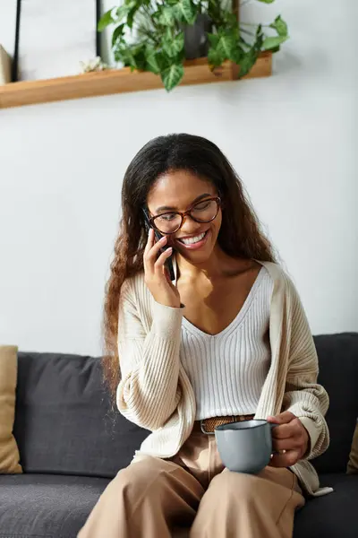 A cheerful woman chats on the phone while relaxing on her sofa, holding a cup of coffee. — Stock Photo