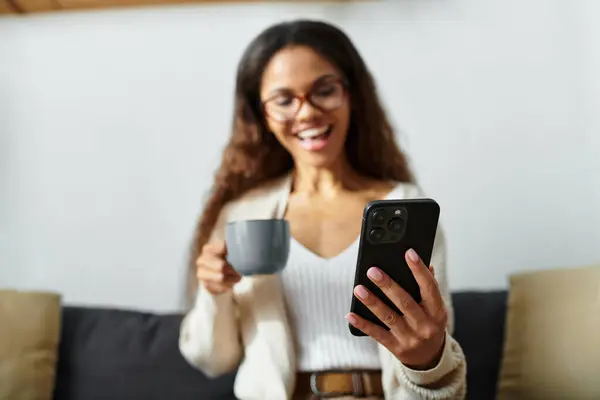 A smiling woman sips coffee and engages with her smartphone in a relaxed setting. — Stock Photo