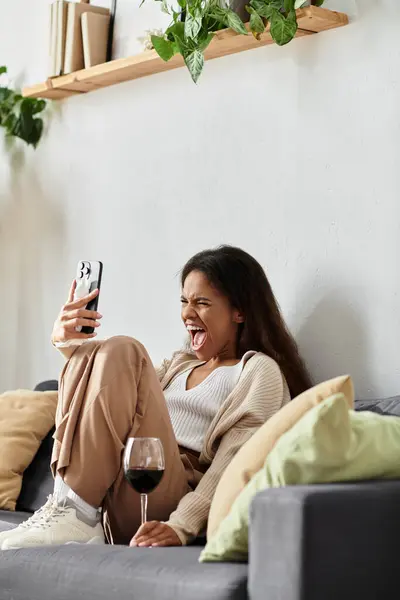 Una giovane donna ride eccitata durante una videochiamata mentre si rilassa su un divano con il suo drink. — Foto stock